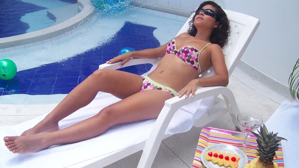 A person relaxing by a poolside lounge chair with a table beside them holding a plate of fruit and a glass.