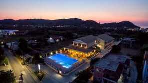 A large building with a swimming pool and multiple balconies, surrounded by hills and other buildings.