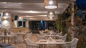 An outdoor dining area with wicker chairs and tables, stone walls, and hanging pendant lights. - Olive Grove Resort (Corfu)