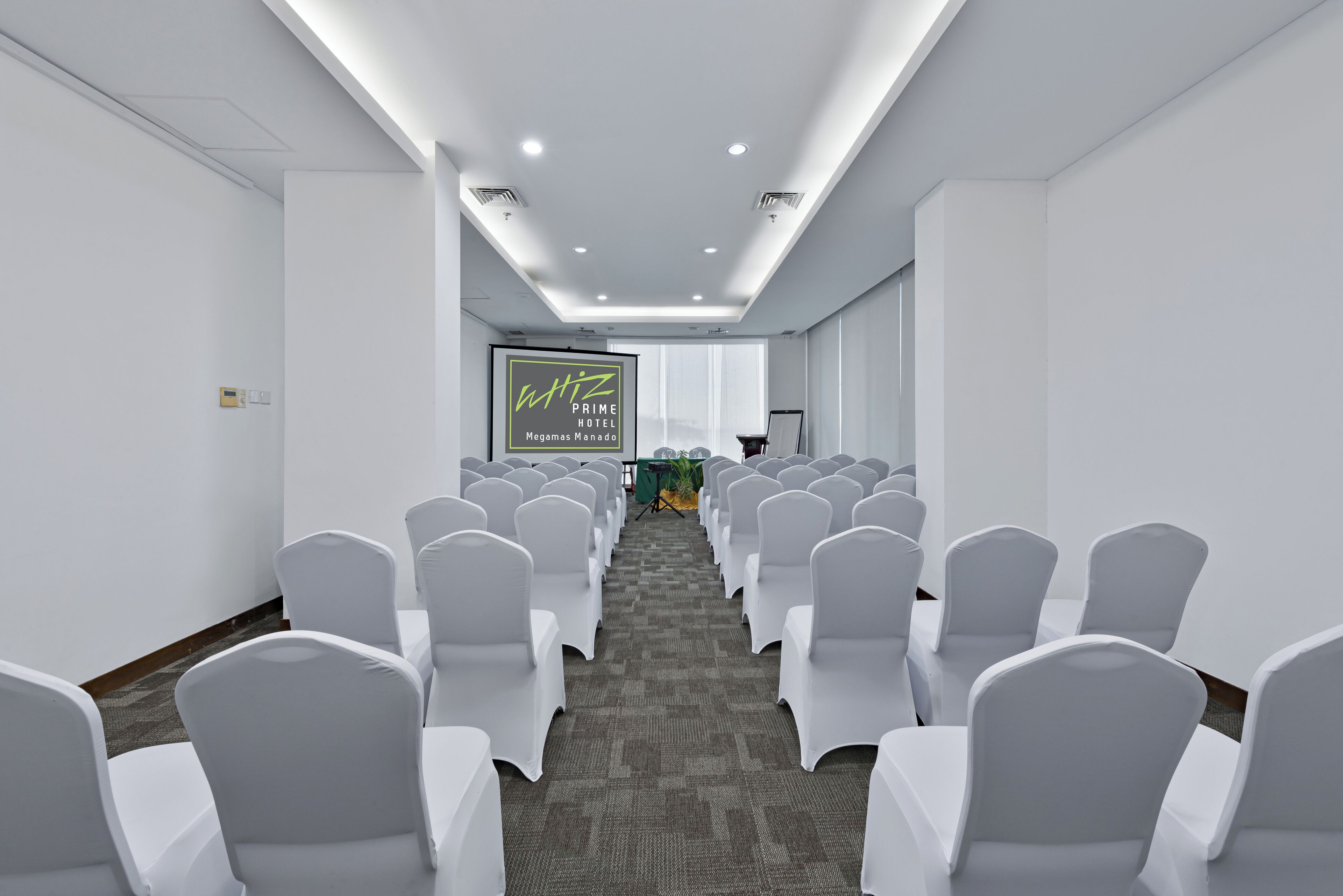 Un salón de conferencias con filas de sillas blancas, una pantalla de proyector y una mesa con mantel verde.