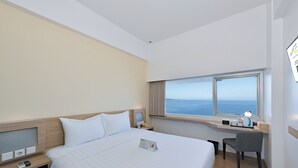 Una camera d'albergo con un letto grande, una scrivania con una sedia, una finestra vista mare e un frigobar.