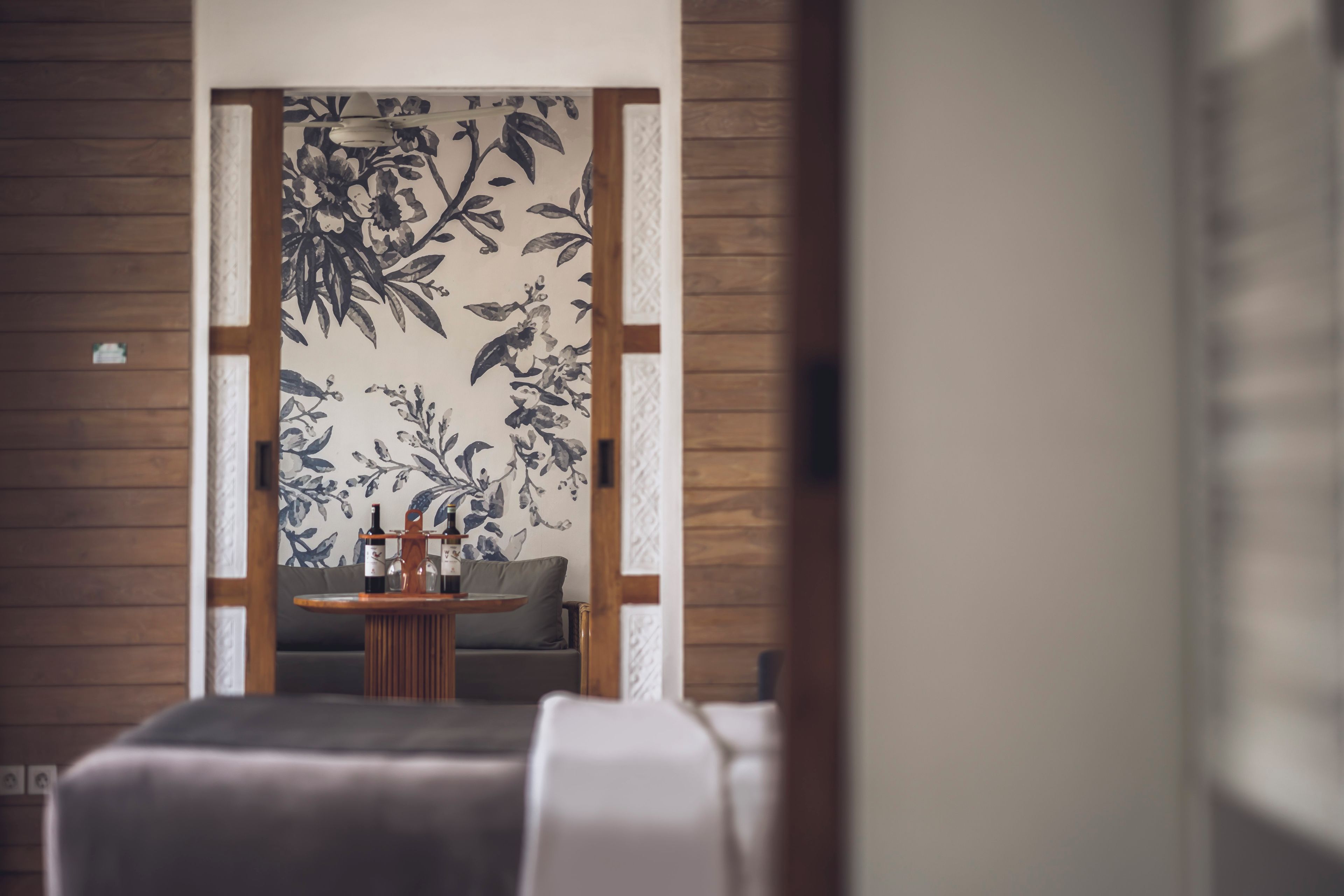 Quarto com parede de madeira, um sofá, uma mesa redonda pequena com uma garrafa e copos, e papel de parede floral.