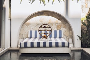 Uma área de estar à beira da piscina com arco de pedra, almofadas listradas e uma pequena mesa com um globo e objetos decorativos.