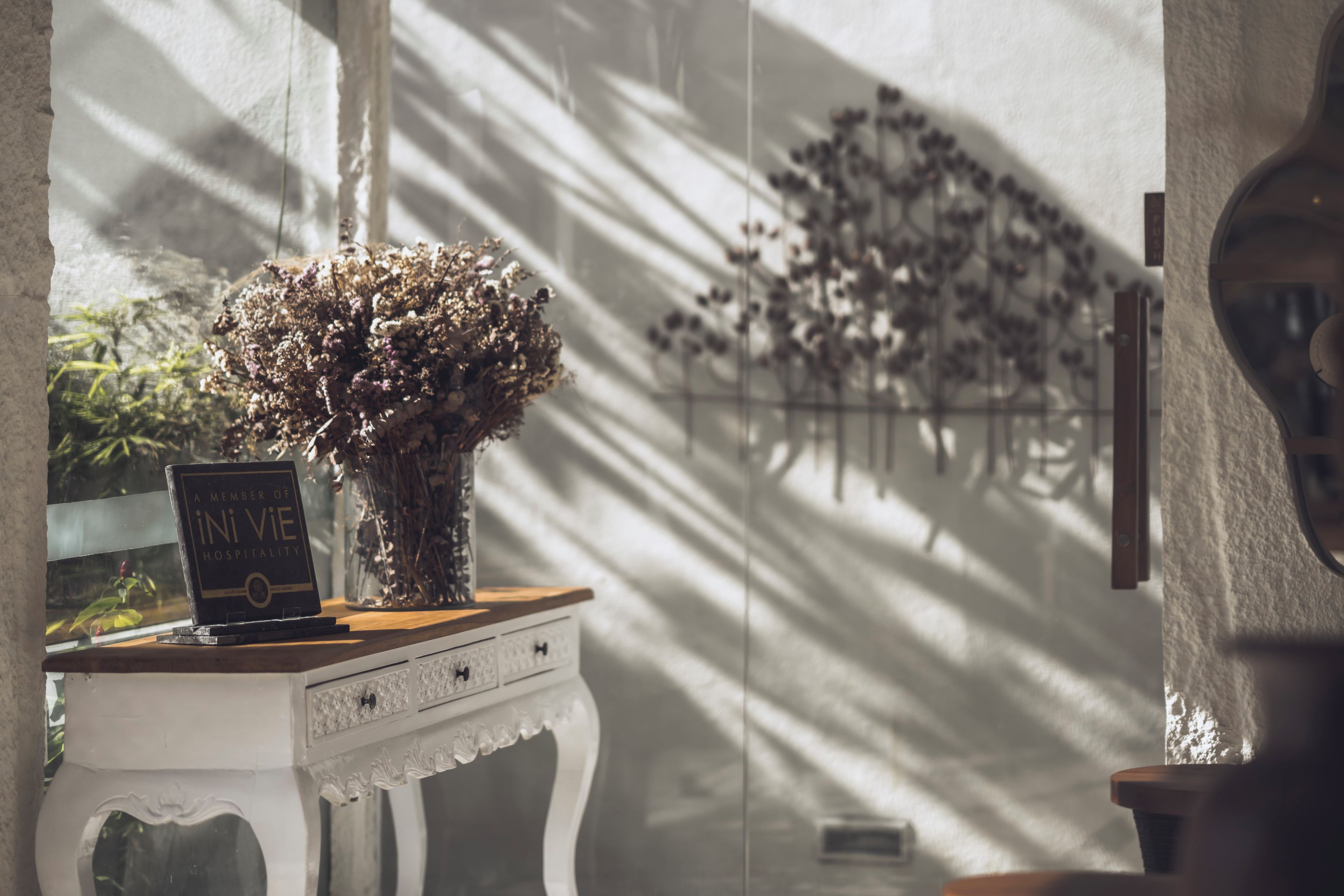 A room with a white table holding a book titled 'A MEMBER OF INI VIE HOSPITALITY' and a vase with dried flowers.