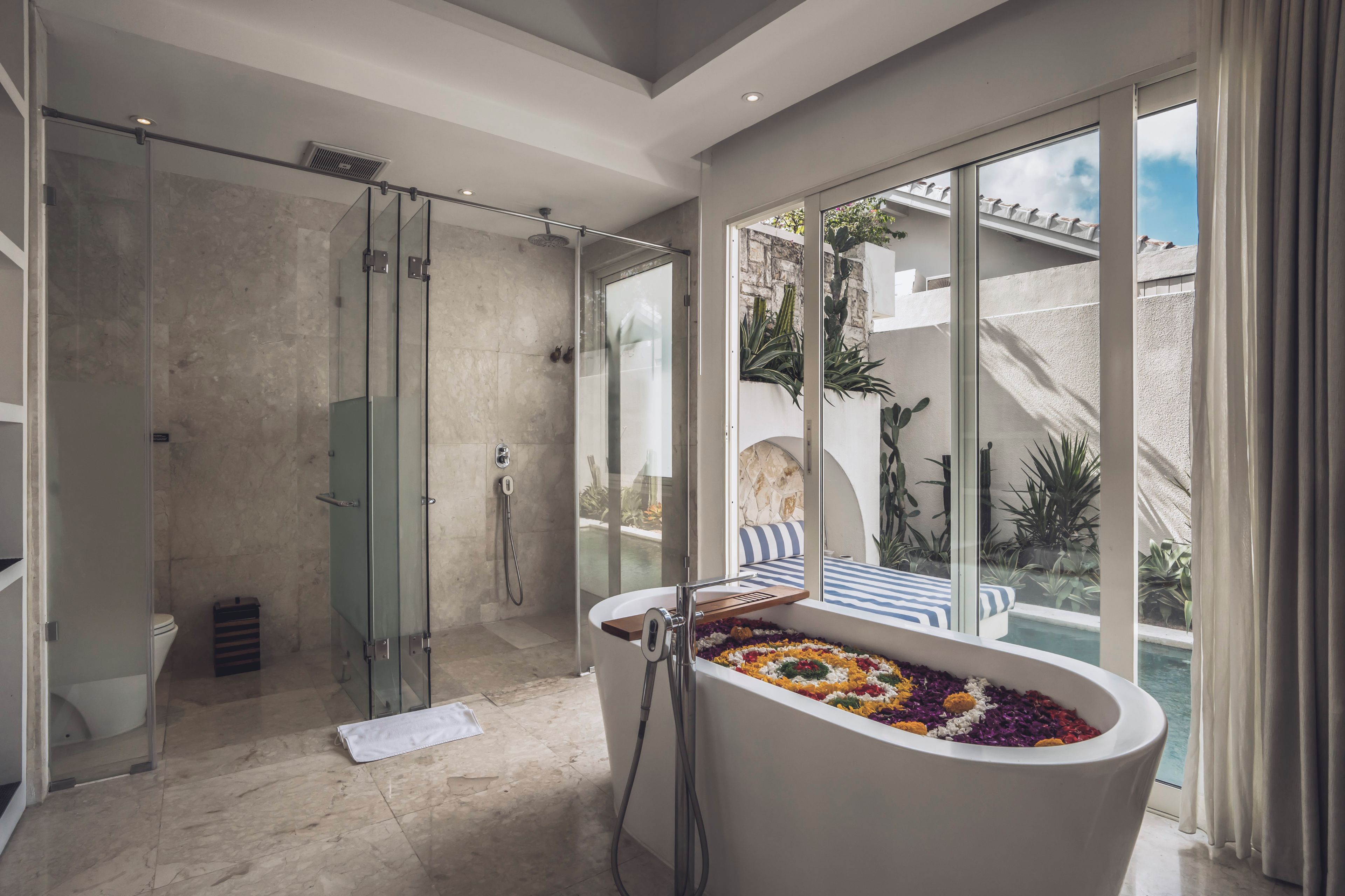 A modern bathroom with a glass-enclosed shower, a freestanding bathtub with colorful bath salts, and a view of a pool and outdoor area through large windows.