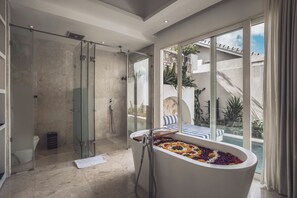 A modern bathroom with a glass-enclosed shower, a freestanding bathtub with colorful bath salts, and a view of a pool and outdoor area through large windows. - Aleva Villa Seminyak by Ini Vie Hospitality (Seminyak)
