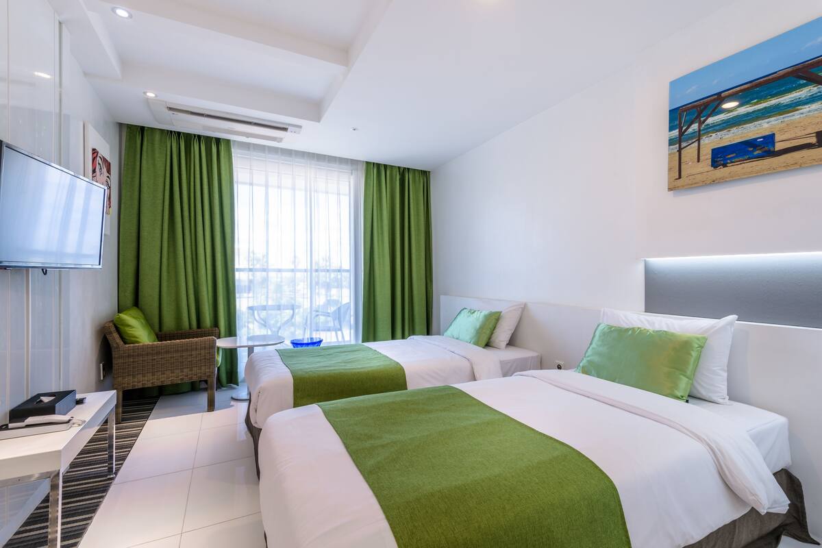 A hotel room with two beds, a TV, a small table, and a window with green curtains.