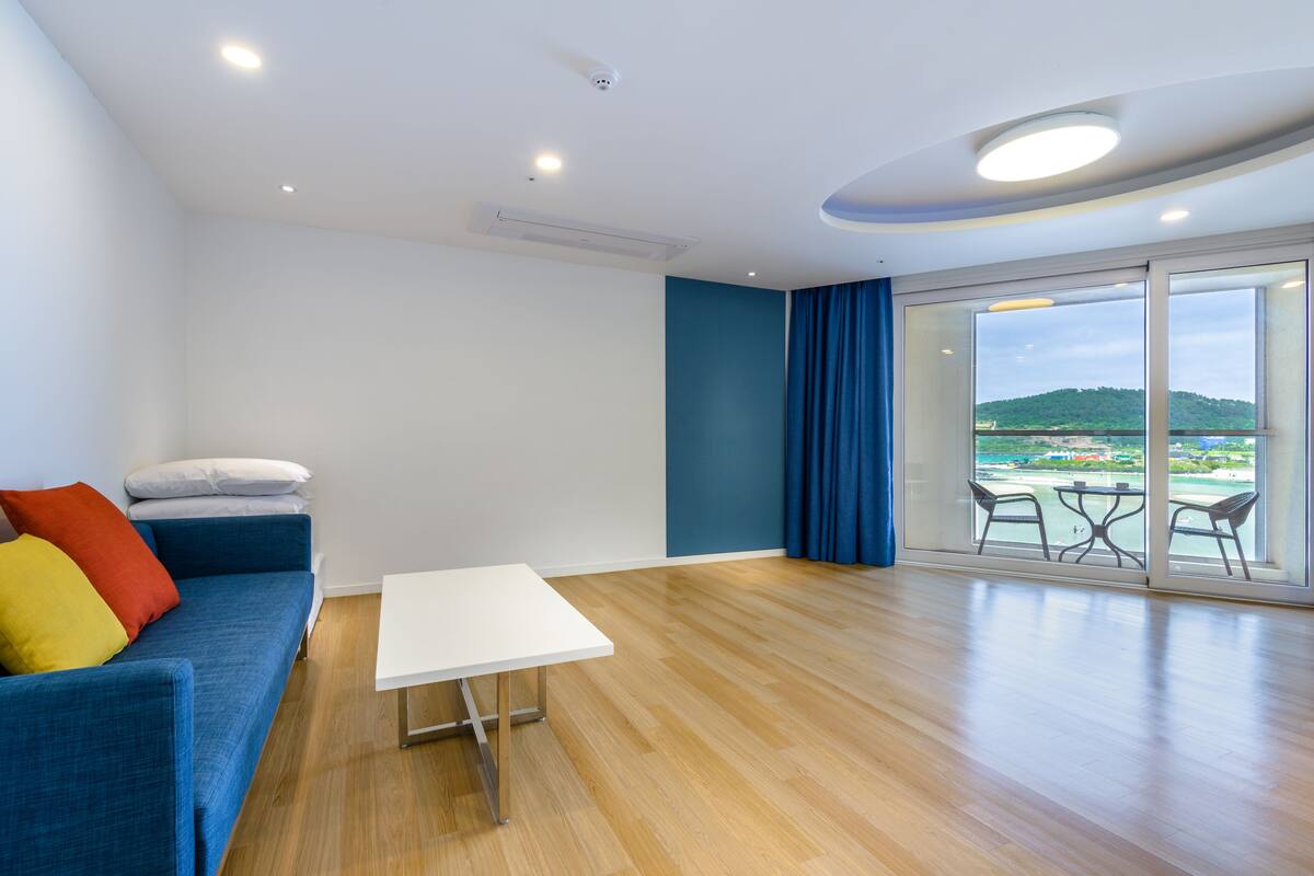 A modern living room with a blue sofa, a white coffee table, and a large sliding glass door leading to a balcony with a view of a green hill.
