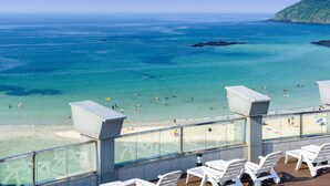 A balcony with white lounge chairs overlooking a beach with people swimming and sunbathing.