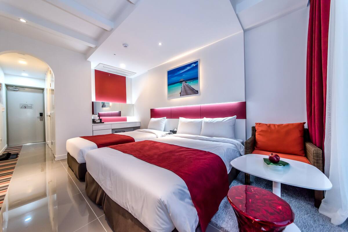 A modern hotel room with a large bed, a separate sofa, a small table, and a red accent wall.