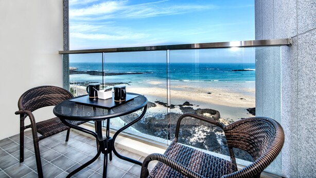 A balcony with wicker chairs and a table, overlooking a beach with clear skies.