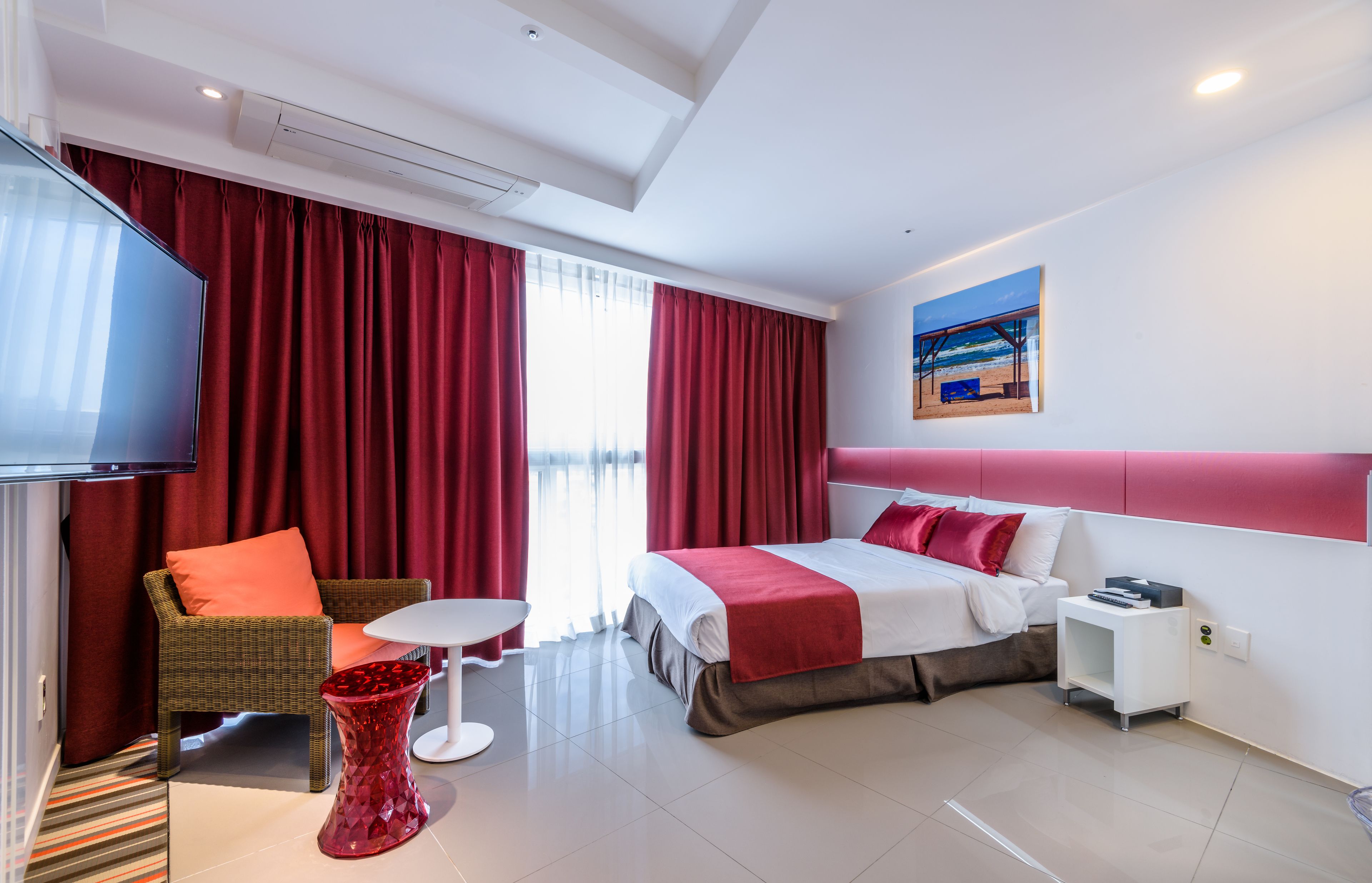 Ein Hotelzimmer mit Bett, Fernseher, rotem Vorhang, Korbsessel, kleinem Tisch und einem Bild an der Wand.