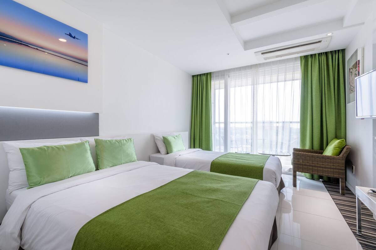 A hotel room with two beds, green bedding, a view of the city, and a large window with green curtains.