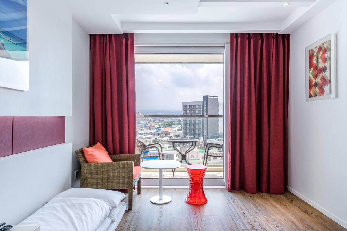 A hotel room with a large window, a bed, a chair, a small table, and a red stool.