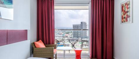 Une chambre d’hôtel avec une grande fenêtre, un lit, une chaise, une petite table et un tabouret rouge.