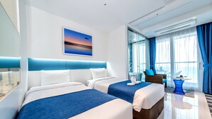 A modern hotel room with two beds, a large window with curtains, a painting on the wall, and a small table with a fruit bowl.