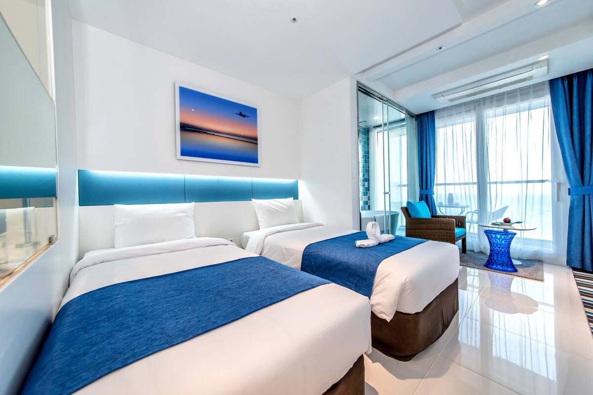 A modern hotel room with two beds, a large window with curtains, a painting on the wall, and a small table with a fruit bowl.