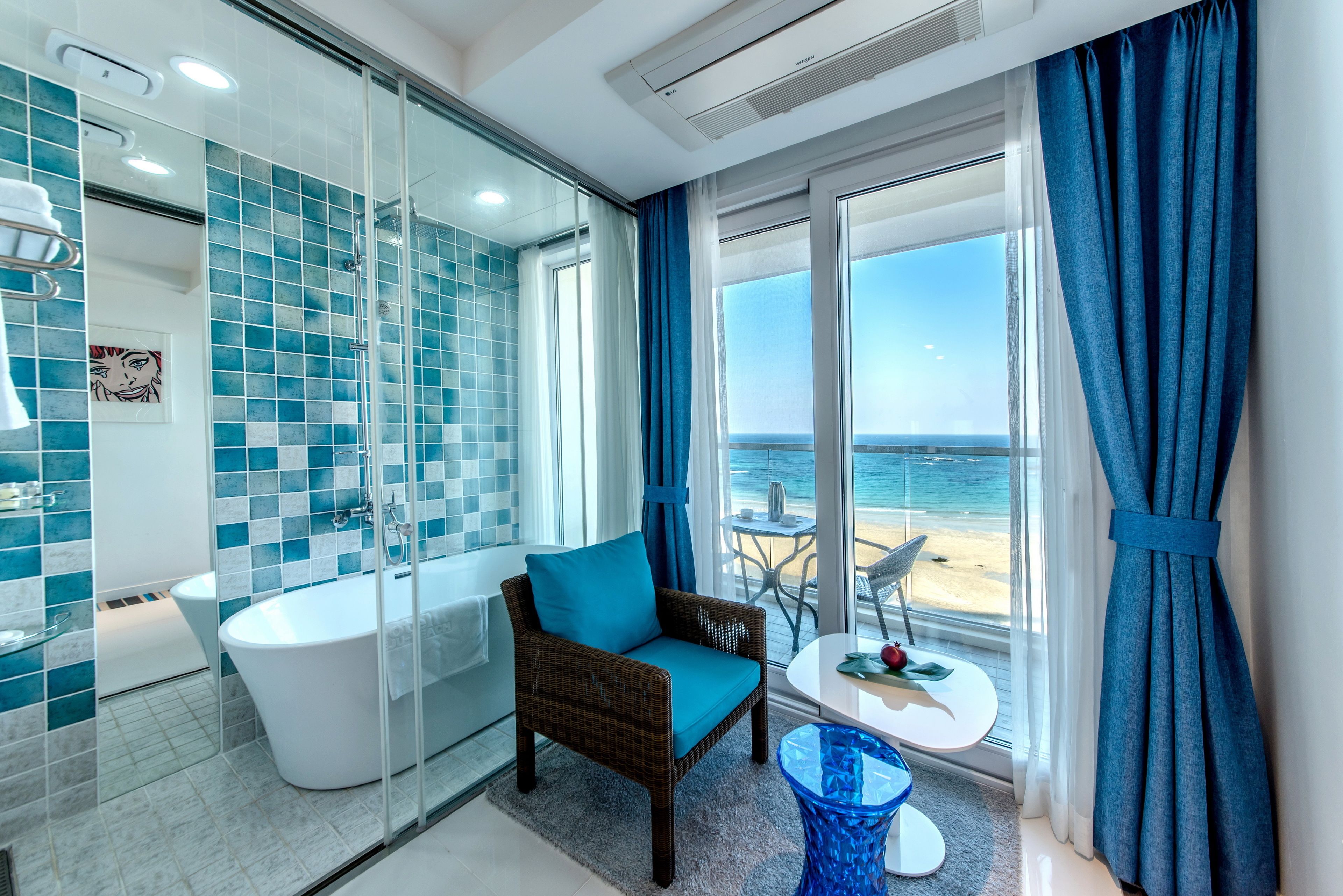 A modern bathroom with a glass-enclosed shower, a white bathtub, and a view of the beach through large windows.