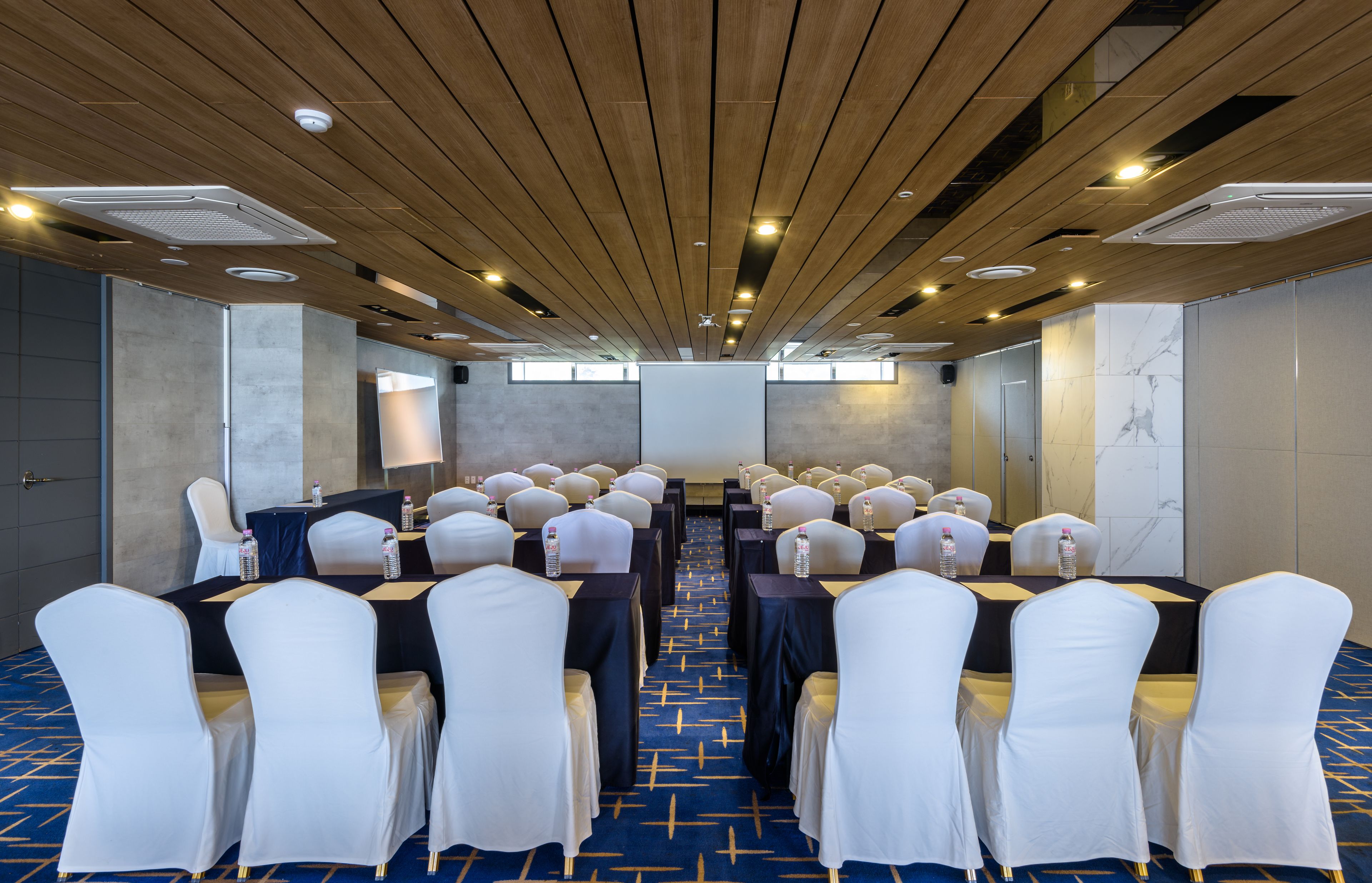 Sala de conferência com fileiras de cadeiras brancas, mesas com garrafas de água e um tapete azul.