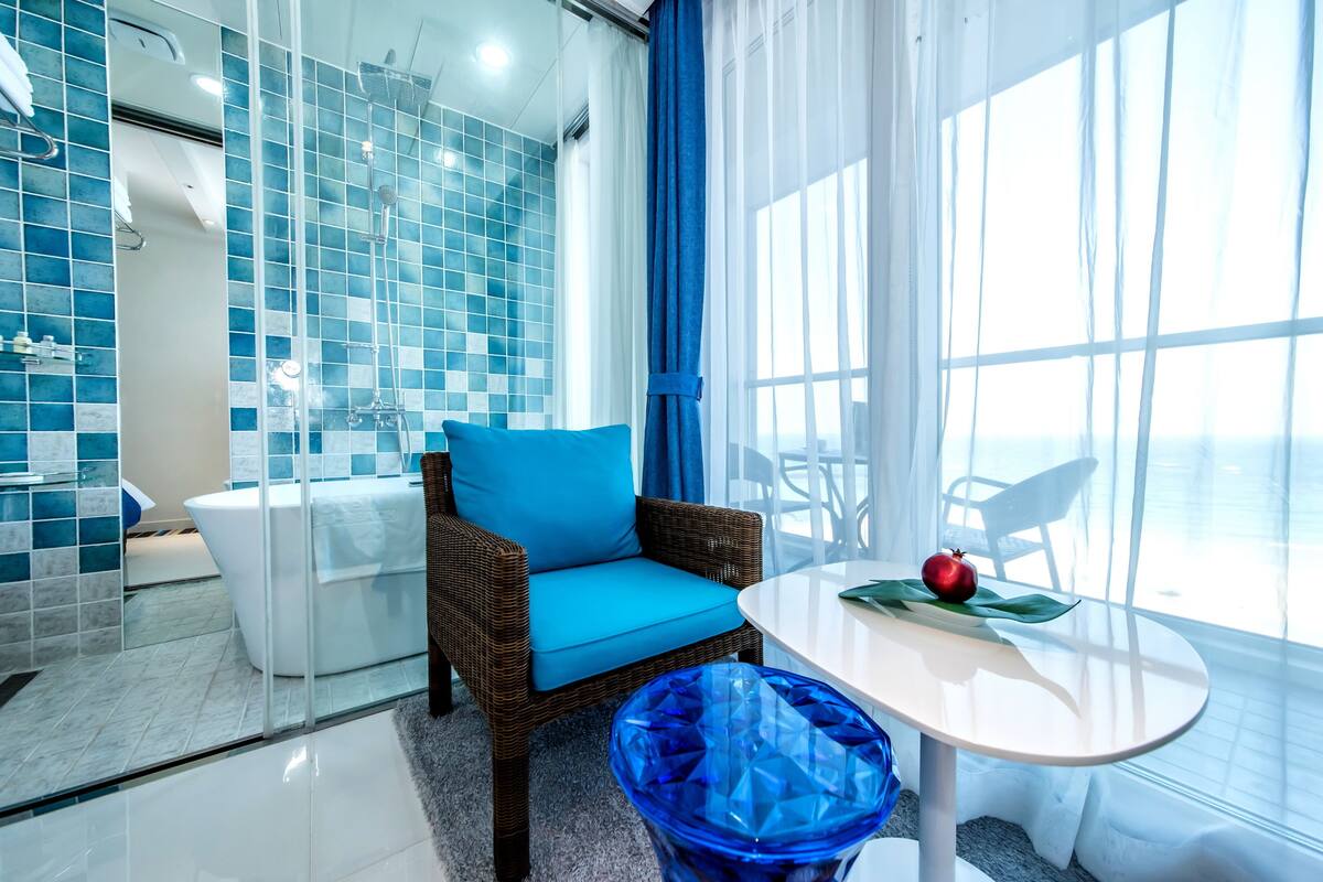 A modern bathroom with blue tiles, a white bathtub, a wicker chair with a blue cushion, and a table with an apple and a green leaf.