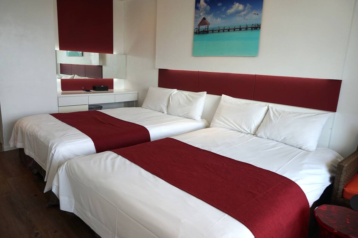A double bed with a red headboard and white bedding, a small bedside table, and a painting of a beach scene.