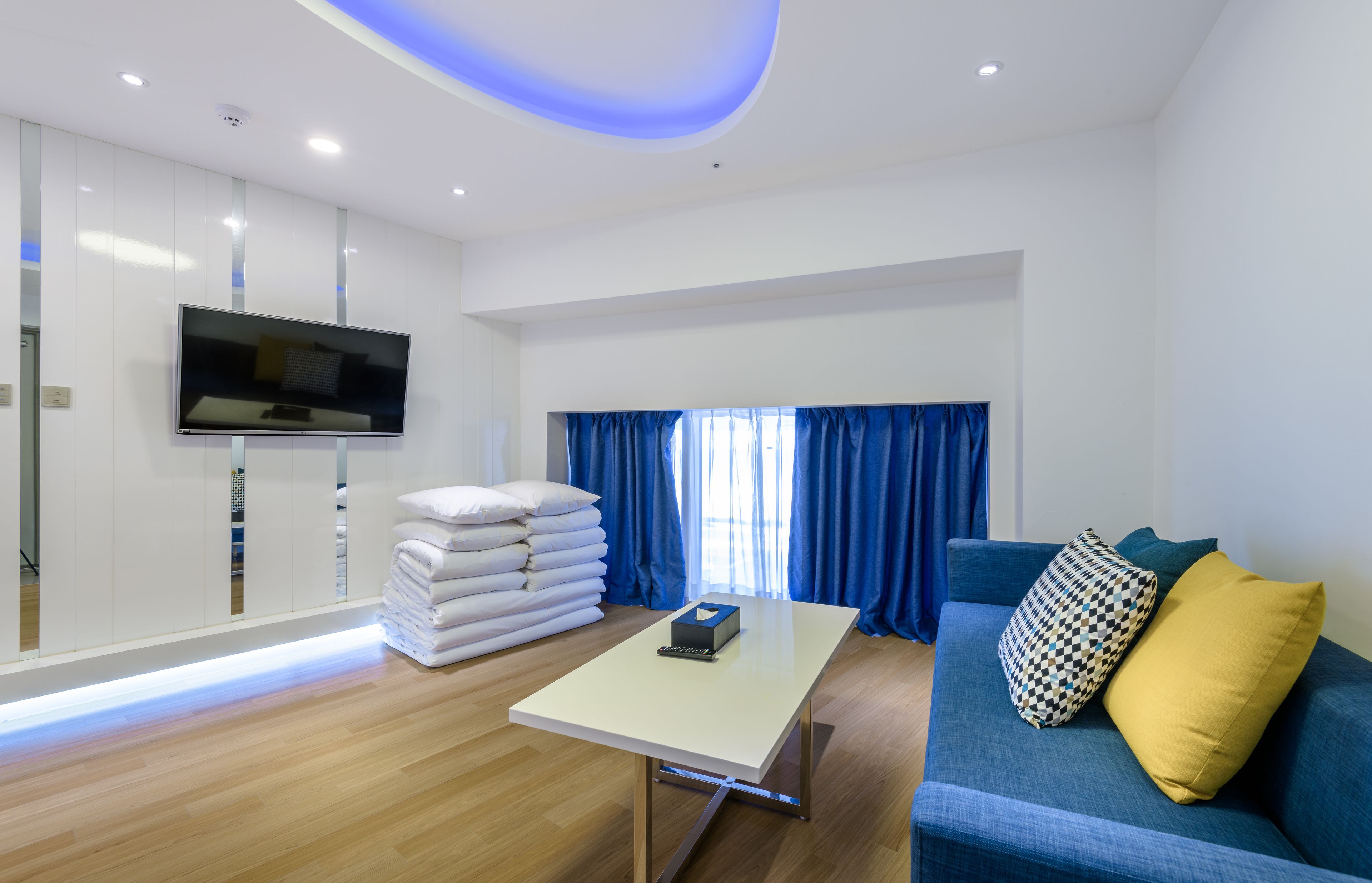 Una habitación de hotel moderna con una televisión de pantalla plana montada en la pared, una mesa blanca con un control remoto, un sofá azul con cojines estampados y una pila de toallas blancas.