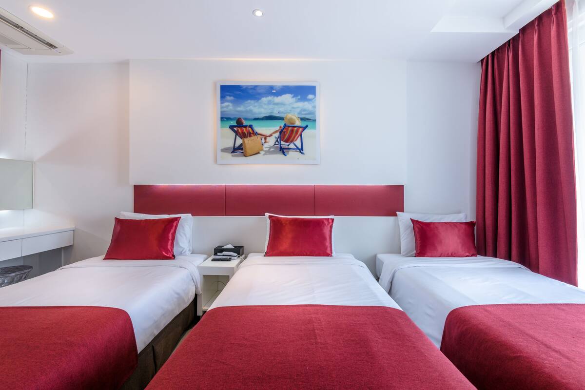 A hotel room with two beds, red bedding, a painting of two people on a beach, and a red curtain.