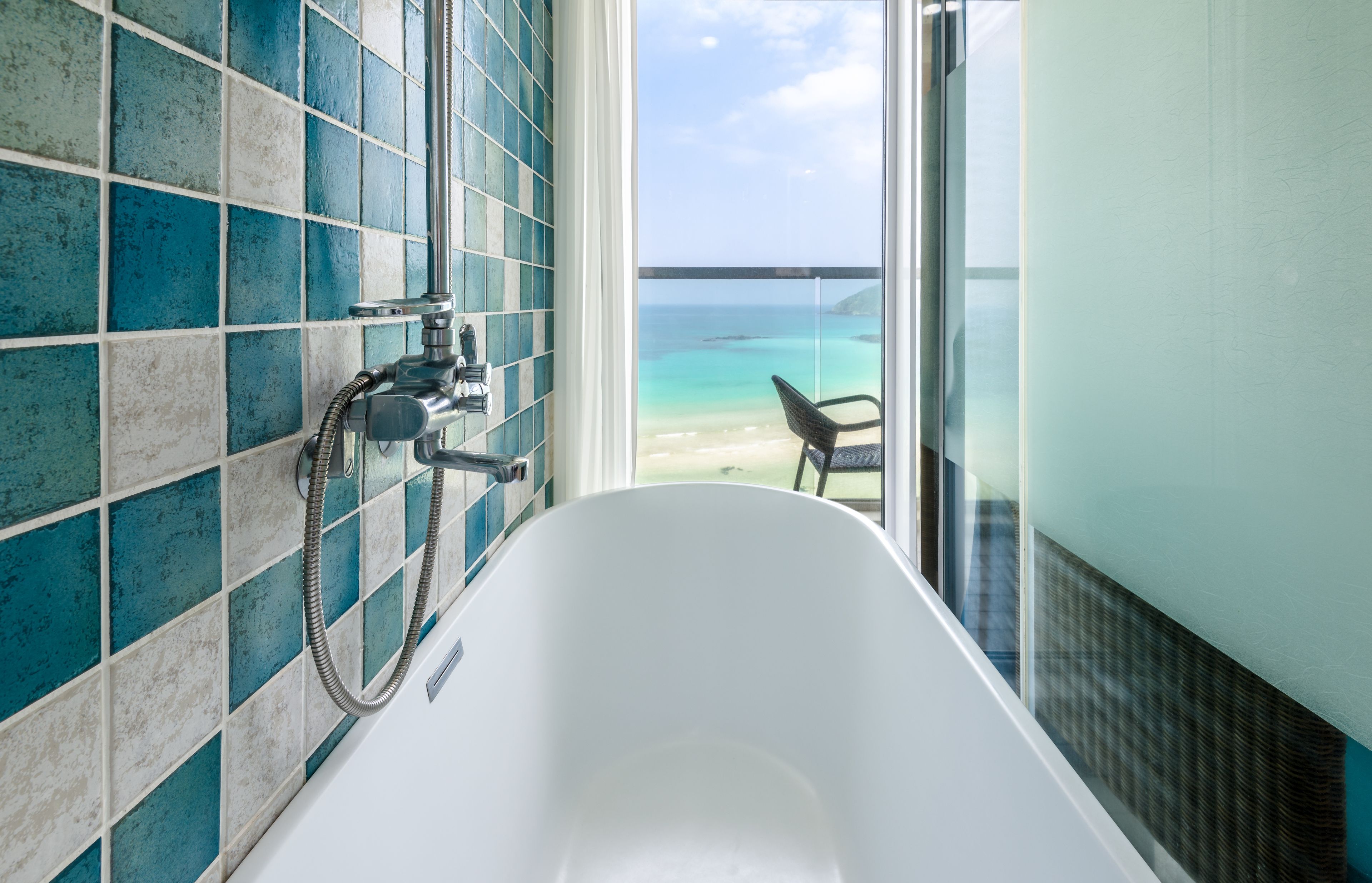 A bathroom with a white bathtub, a shower, and a view of a beach through a glass door.
