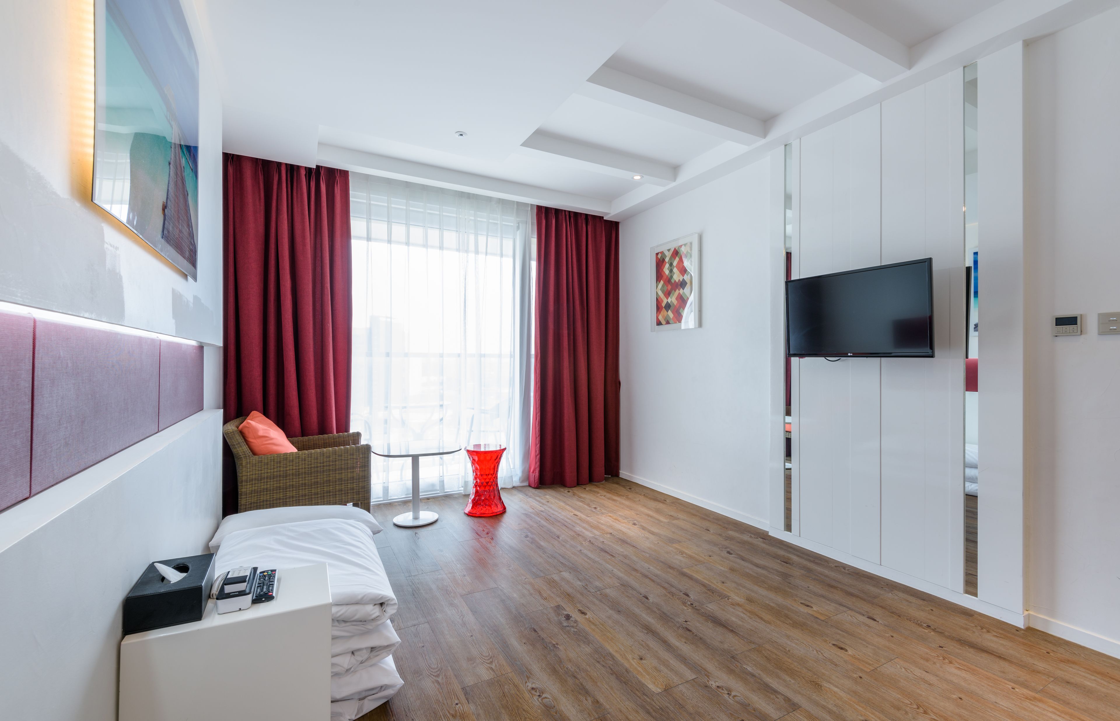 Una habitación de hotel moderna con suelo de madera, una cama con sábanas blancas, una silla roja, una mesita pequeña roja, un televisor de pantalla plana montado en la pared y una ventana con cortinas rojas.