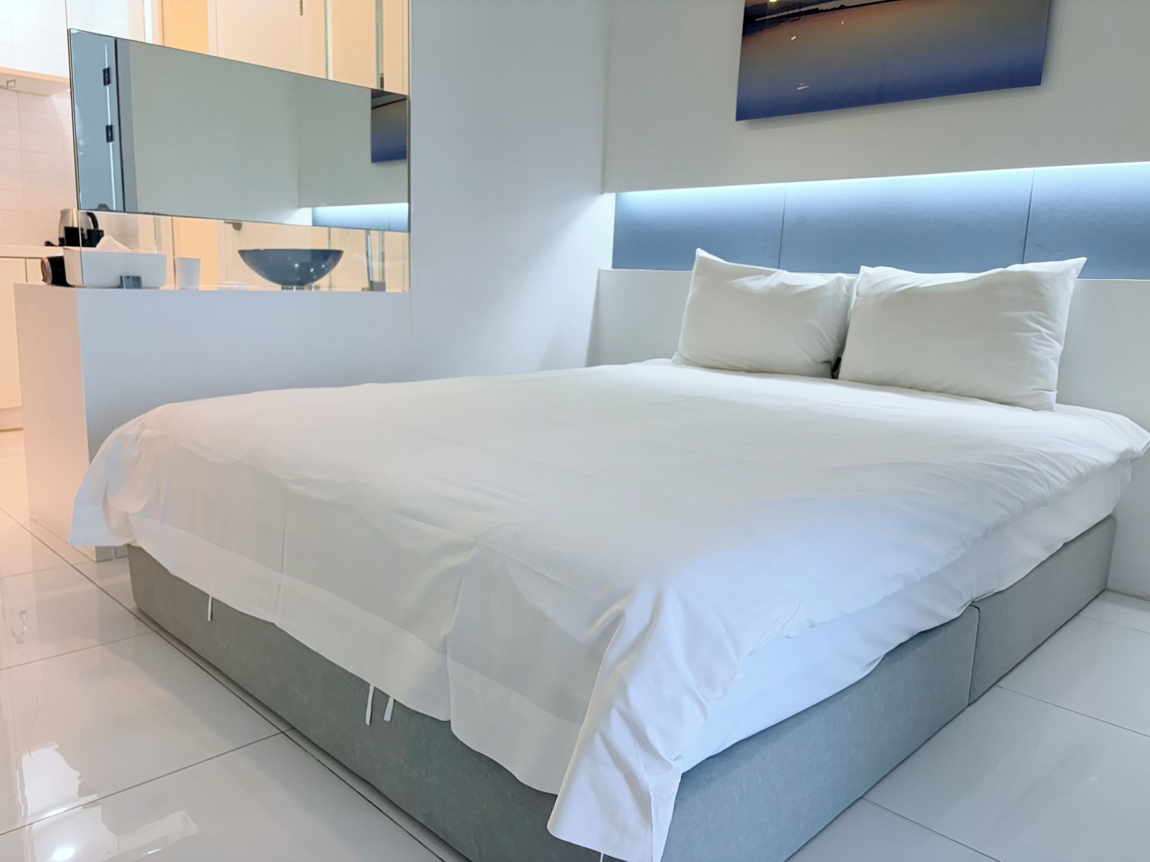 A modern bedroom with a large bed, a headboard with integrated lighting, a flat-screen TV, and a minimalist decor.