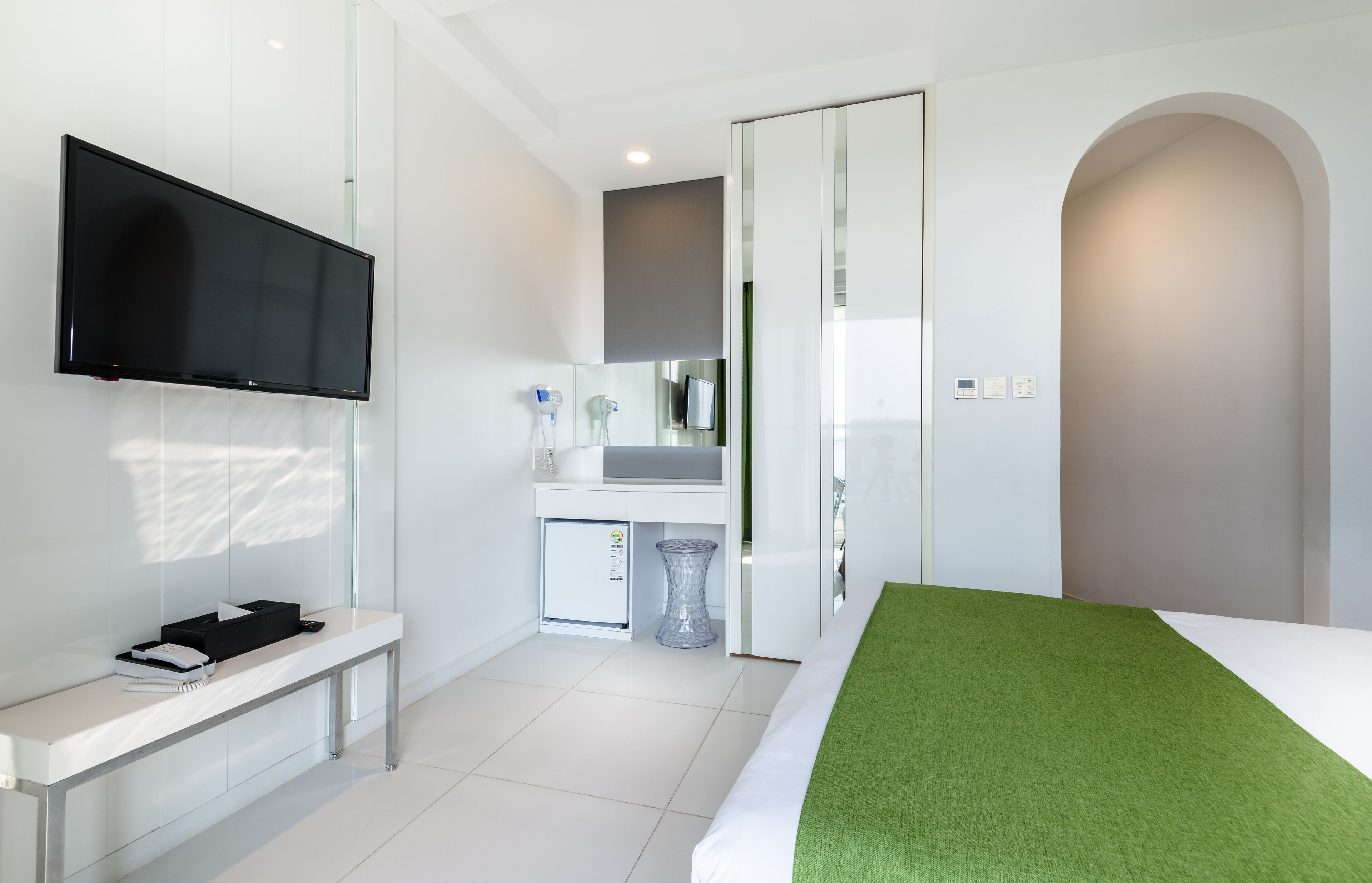 Habitación de hotel moderna con televisión de pantalla plana, una kitchenette pequeña, un espejo y una cama con detalles verdes.