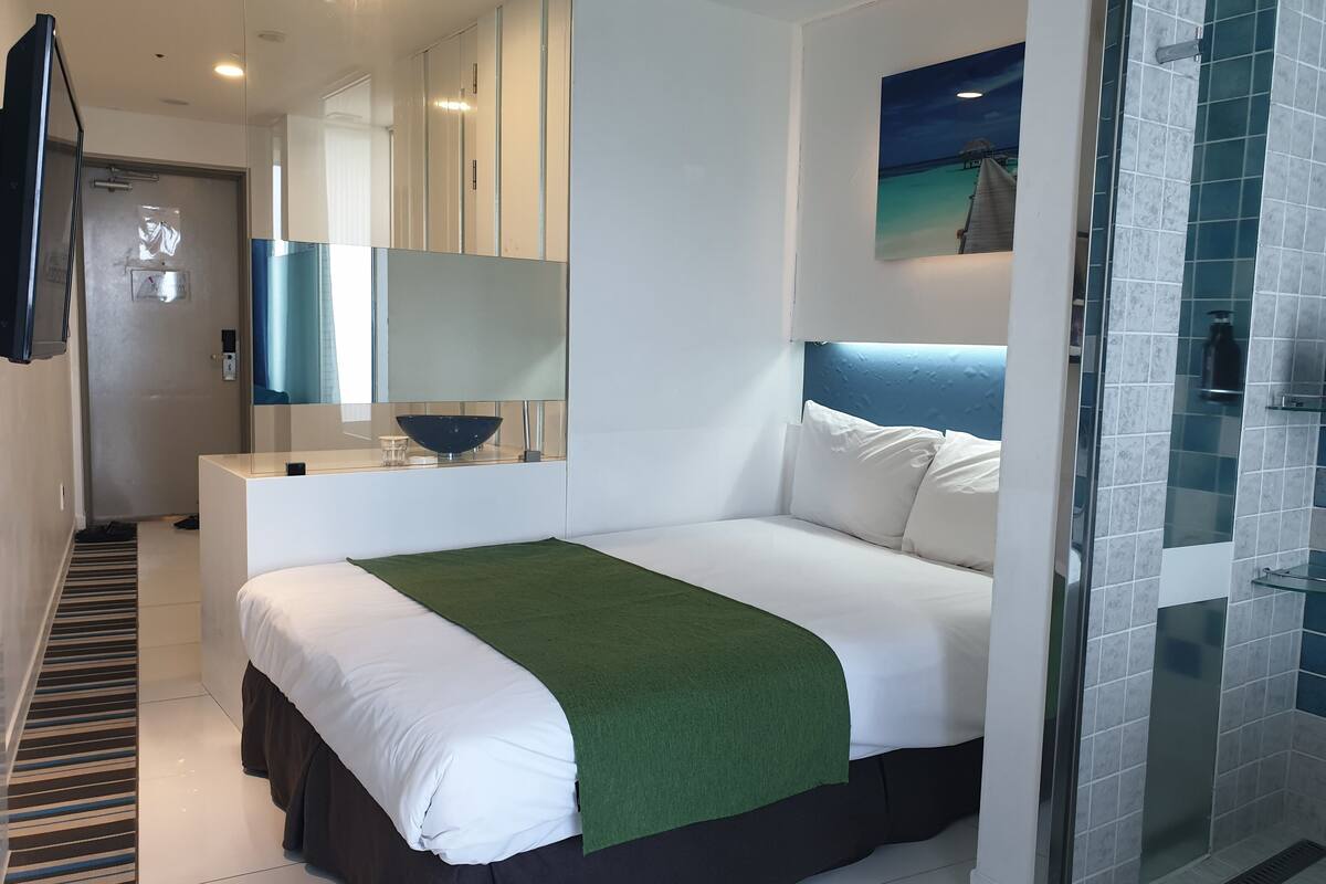 A modern hotel room with a bed, a large mirror, a small bedside table, a green throw blanket, and a shower area.
