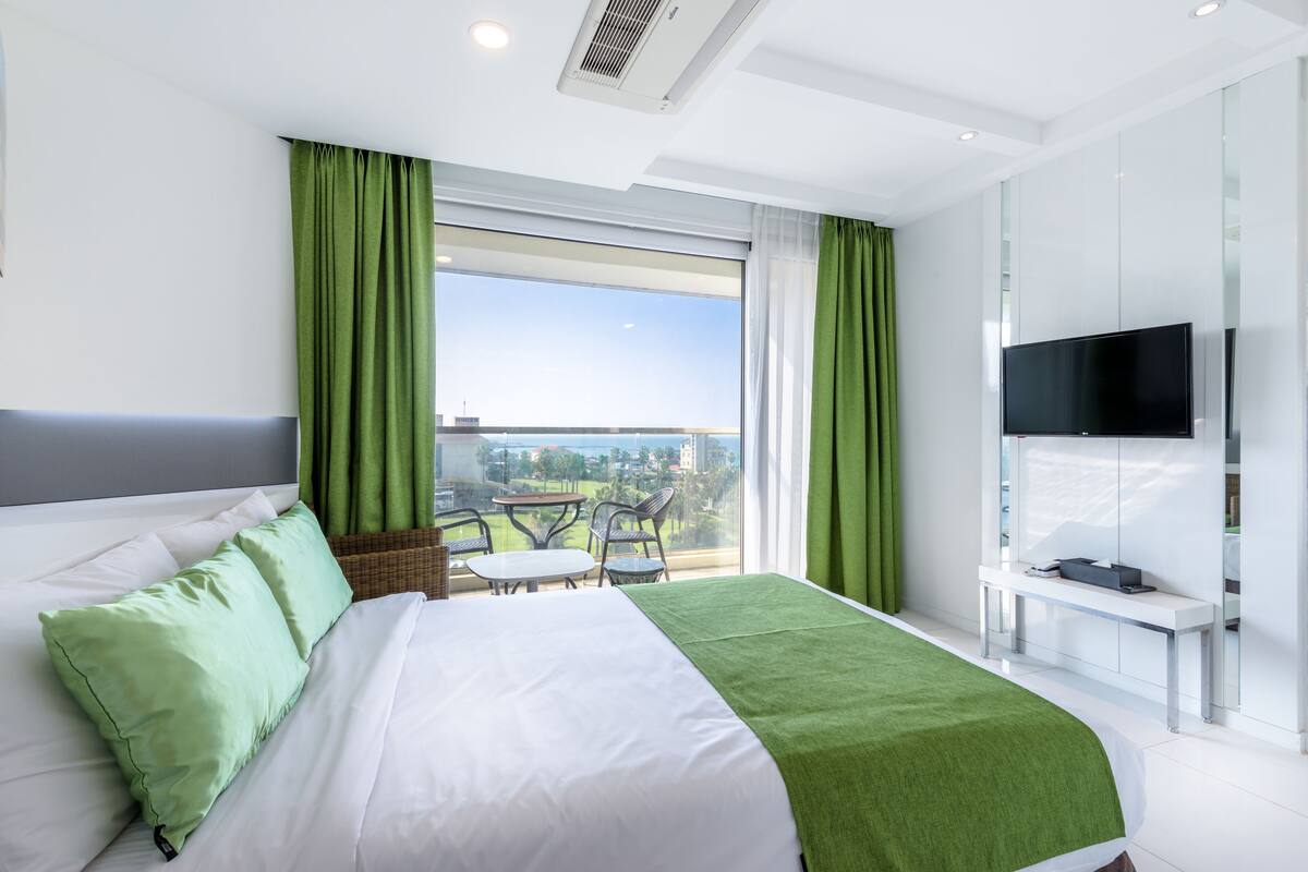 A modern hotel room with a large bed, green curtains, a balcony with a table and chairs, and a TV on a stand.