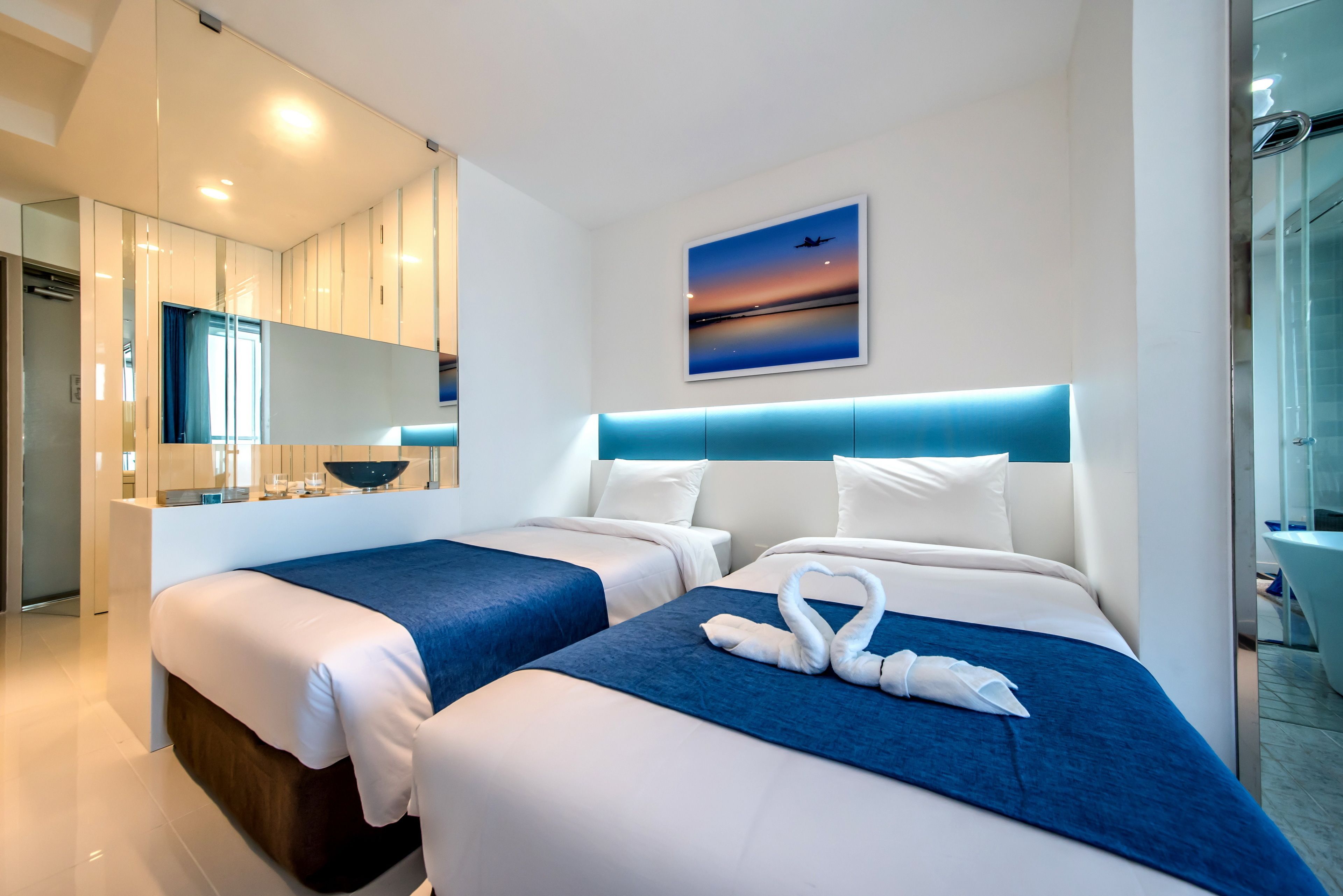 Une chambre d’hôtel moderne avec deux lits, un grand miroir, un tableau représentant un coucher de soleil et une tête de lit bleue.