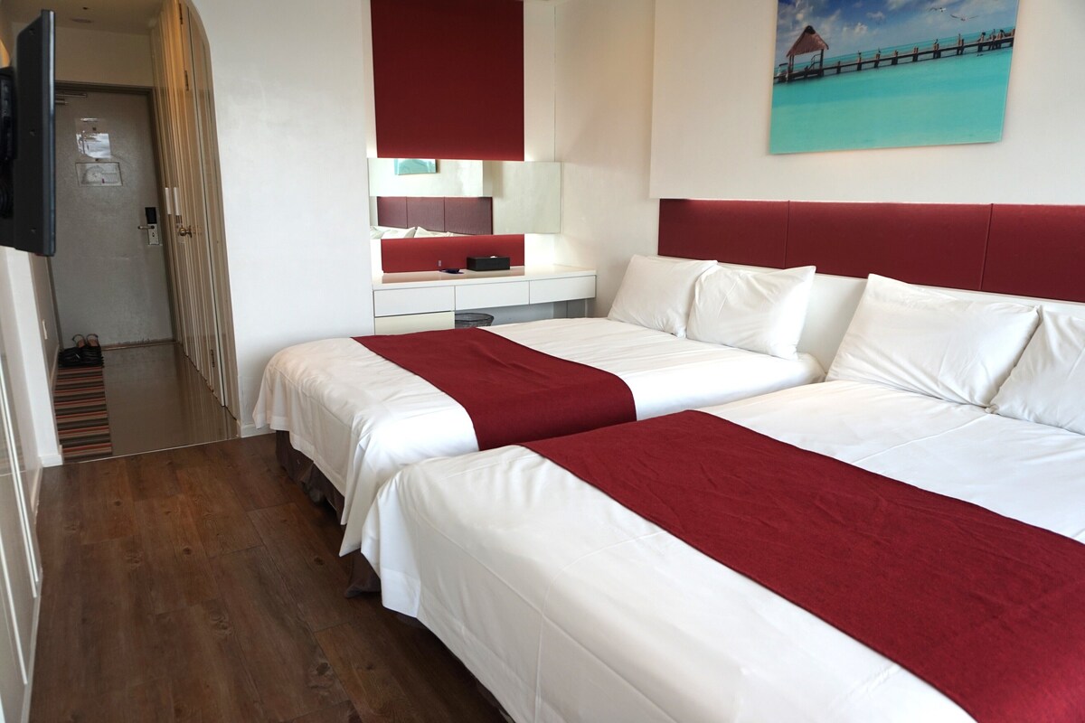Habitación de hotel con dos camas, cabecera roja, mesita de noche blanca, un espejo y un cuadro con una playa.