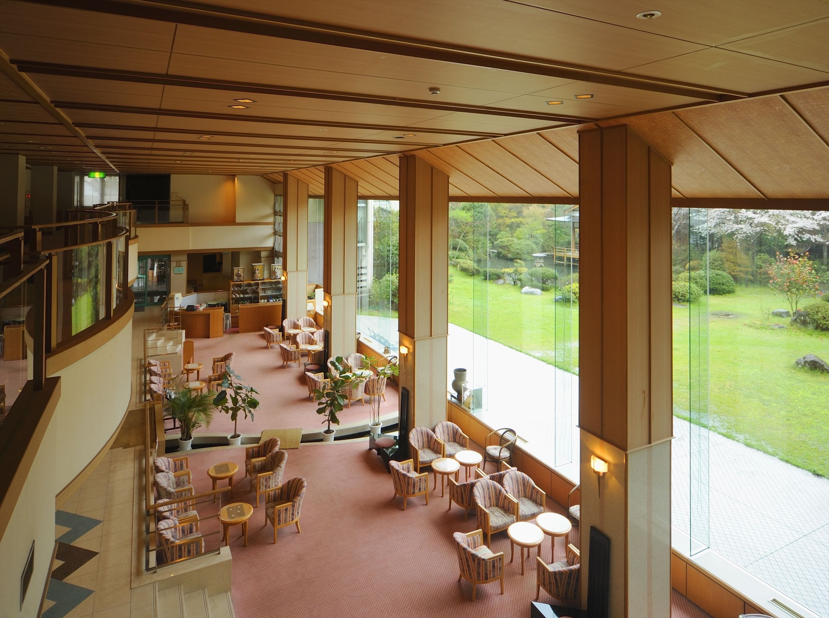 A spacious lobby with a high ceiling, large windows offering a view of a garden, and multiple seating areas with tables and chairs.