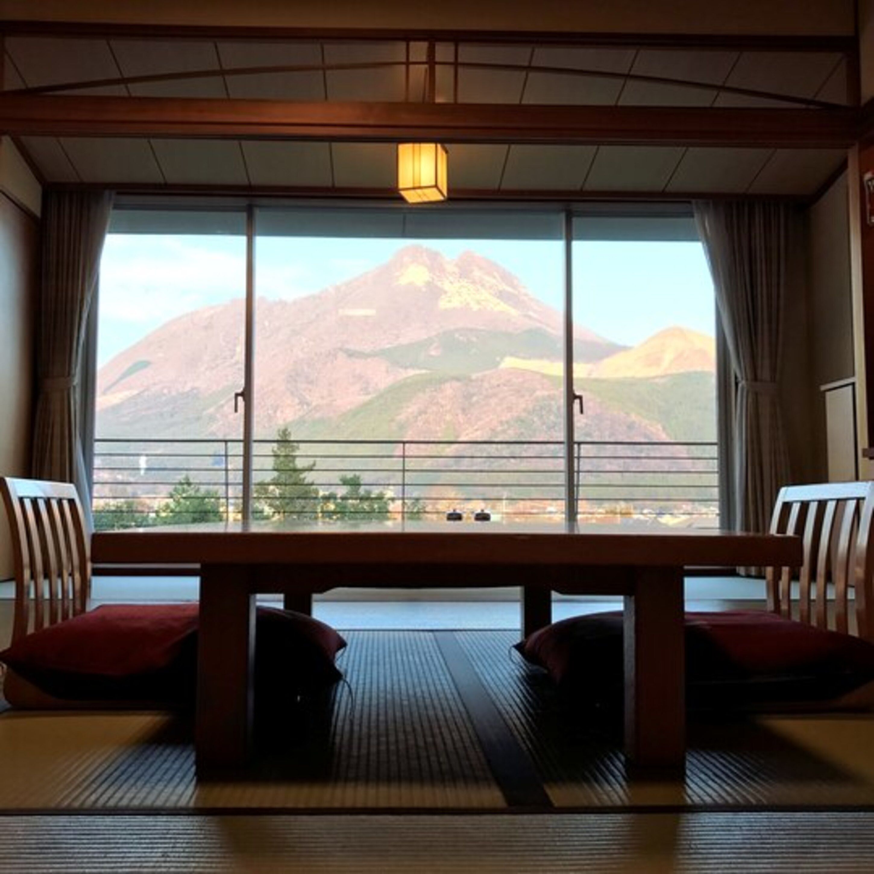 A room with a large window offering a mountain view. There is a wooden table with two chairs and a small table lamp.