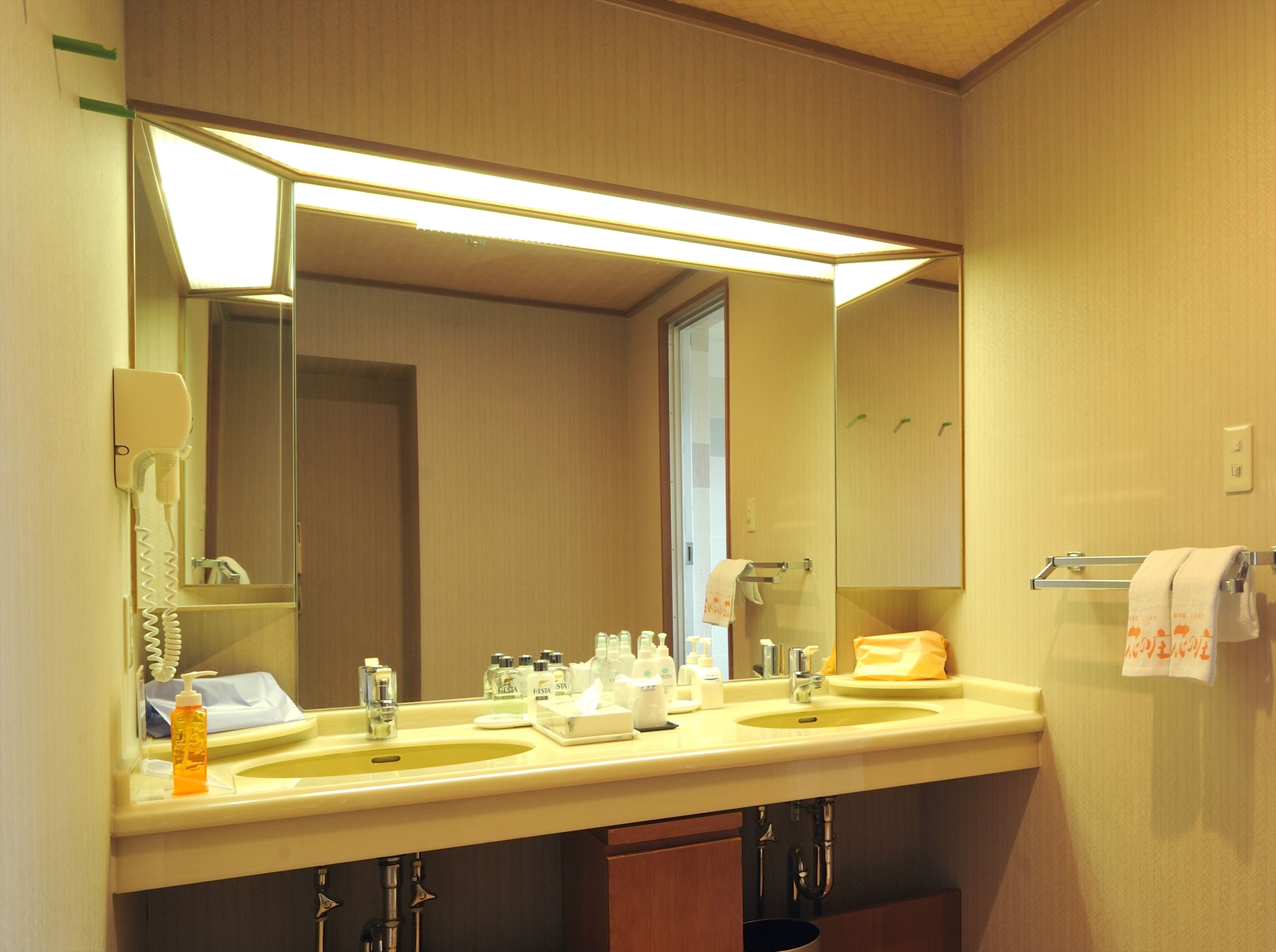 A hotel bathroom with a large mirror, two sinks, a hairdryer, and a towel rack.
