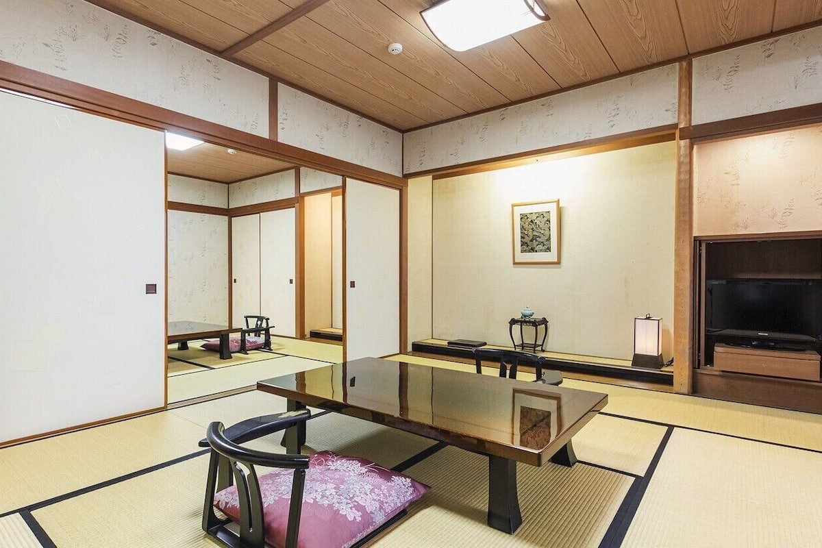 A traditional Japanese room with tatami flooring, a low table, a chair, a television, and a framed picture on the wall.