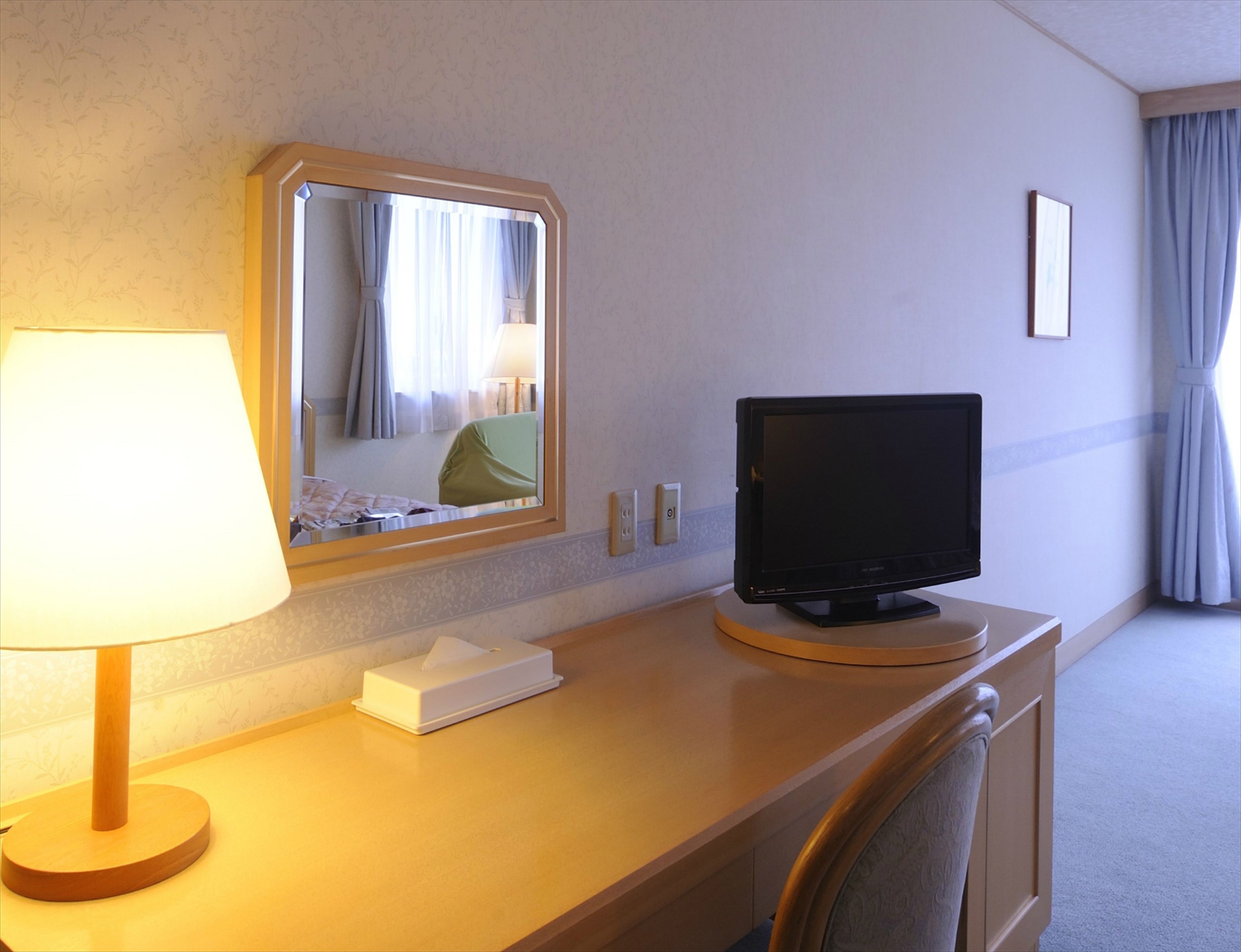 A hotel room with a desk, a lamp, a television, a mirror, and a window with curtains.