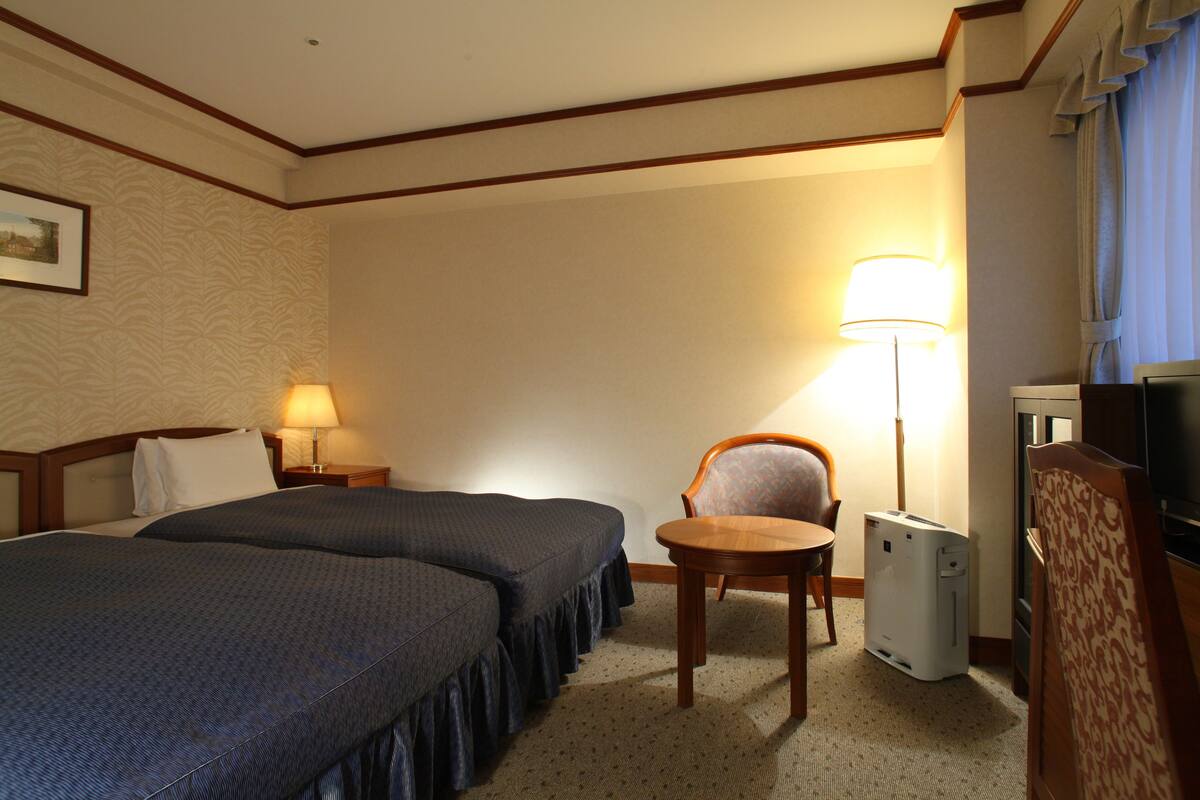A hotel room with two beds, a desk, a chair, a lamp, and a TV.
