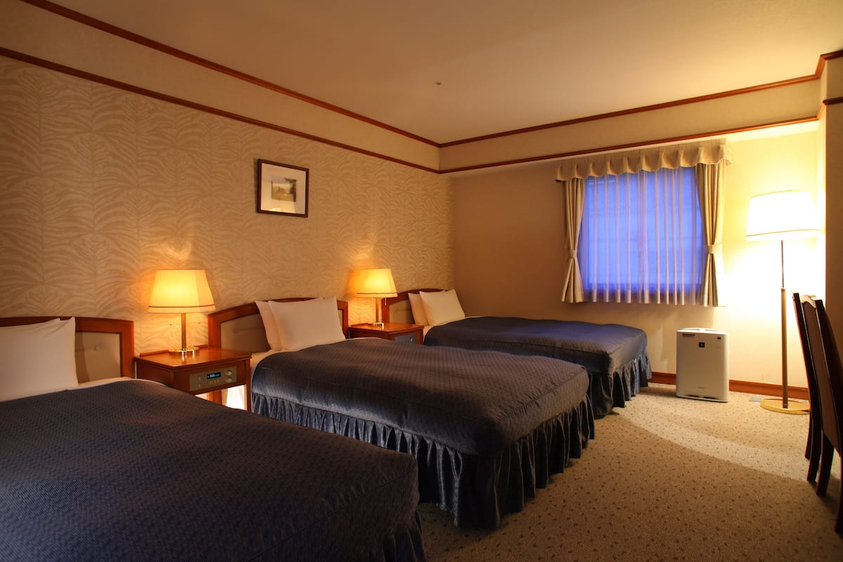 A hotel room with three beds, two bedside tables with lamps, a large window with curtains, and a standing heater.