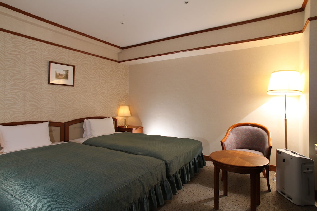 A hotel room with two beds, a chair, a lamp, and a heater.