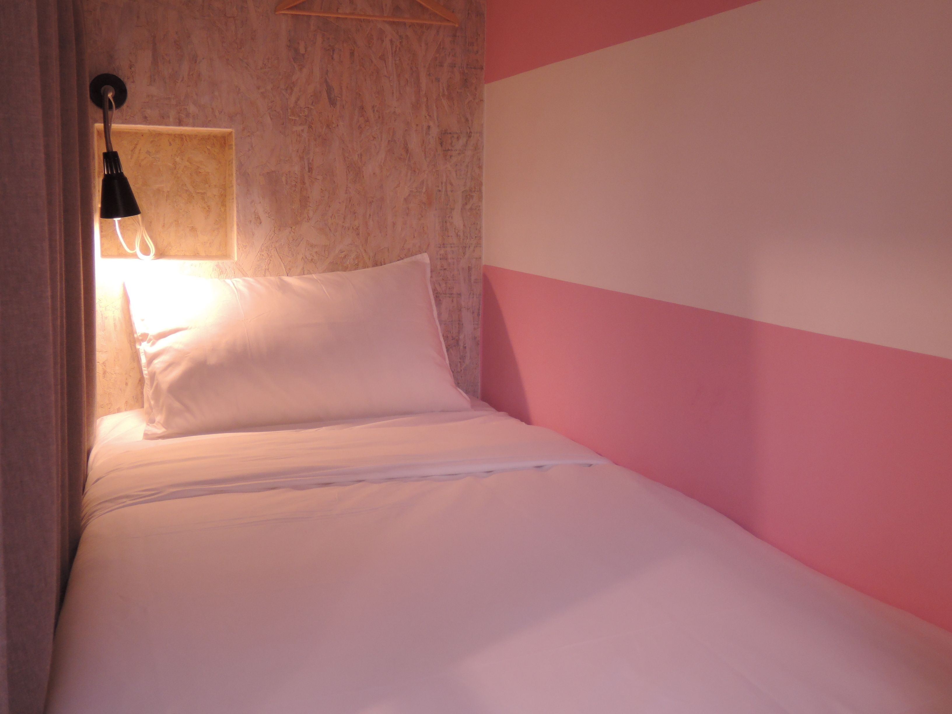 Una cama individual con ropa de cama blanca, una lámpara colgante y una pared rosa y blanca.
