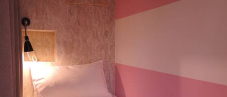 Una cama individual con ropa de cama blanca, una lámpara colgante y una pared rosa y blanca.