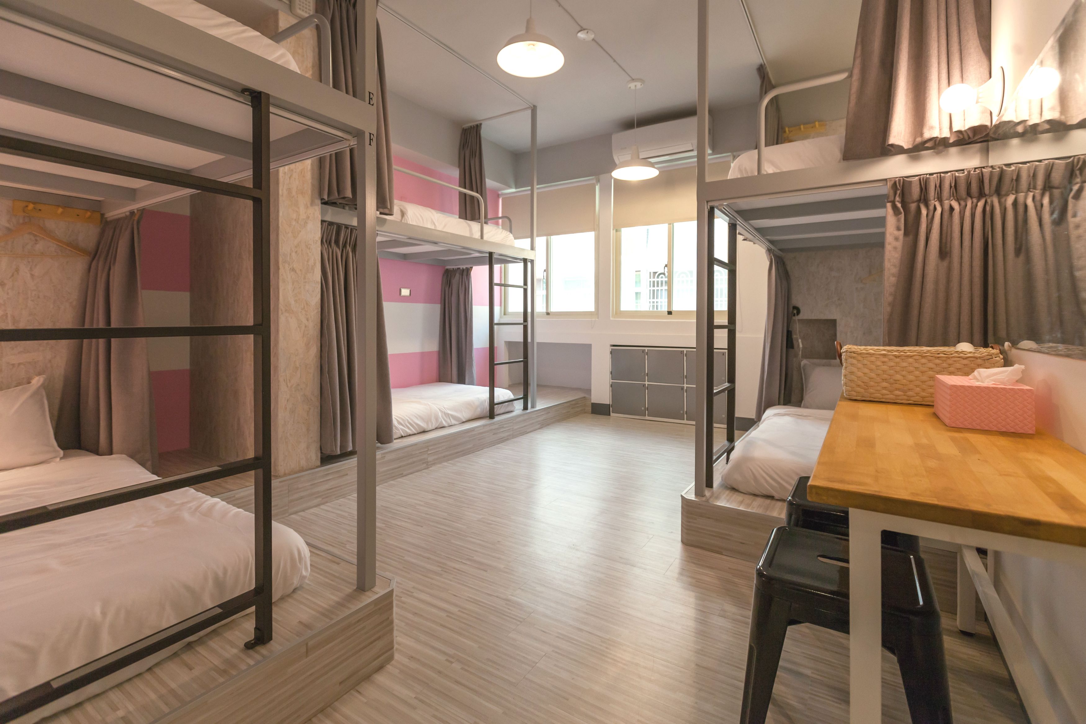 A dormitory room with bunk beds, a wooden table, and a window with curtains.