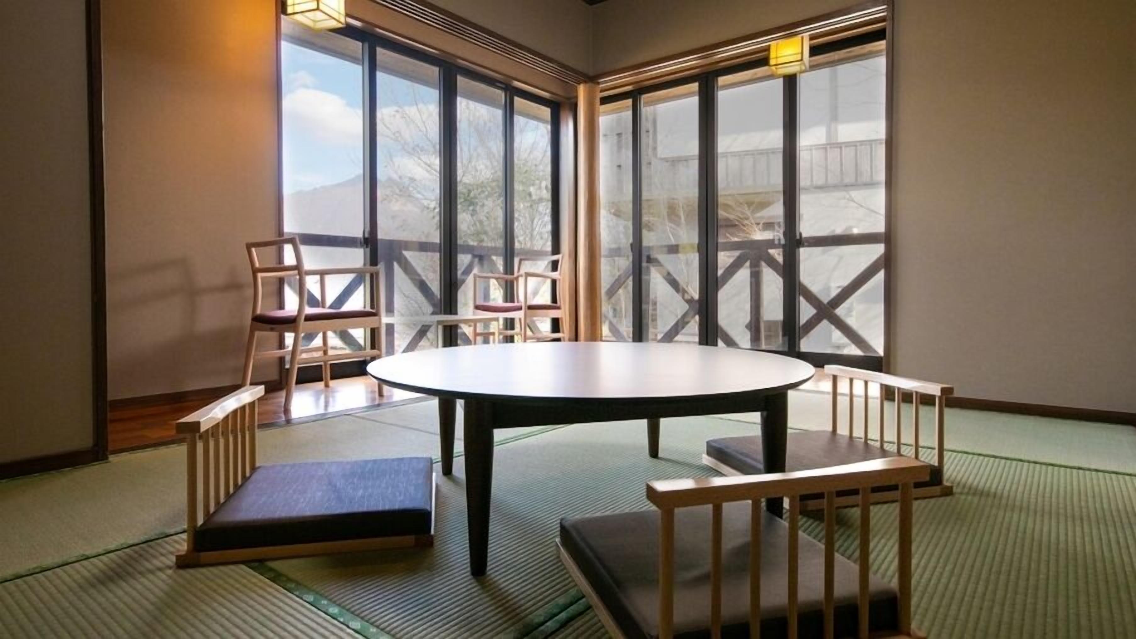 Une pièce avec un sol recouvert de tatamis, une table ronde et des bancs, de grandes fenêtres offrant une vue sur les montagnes.