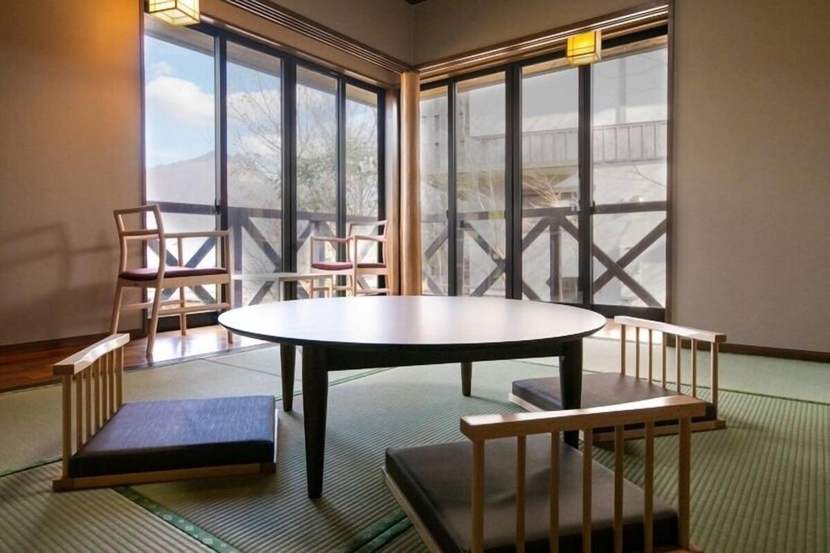 Une pièce avec un sol recouvert de tatamis, une table ronde et des bancs, de grandes fenêtres offrant une vue sur les montagnes.