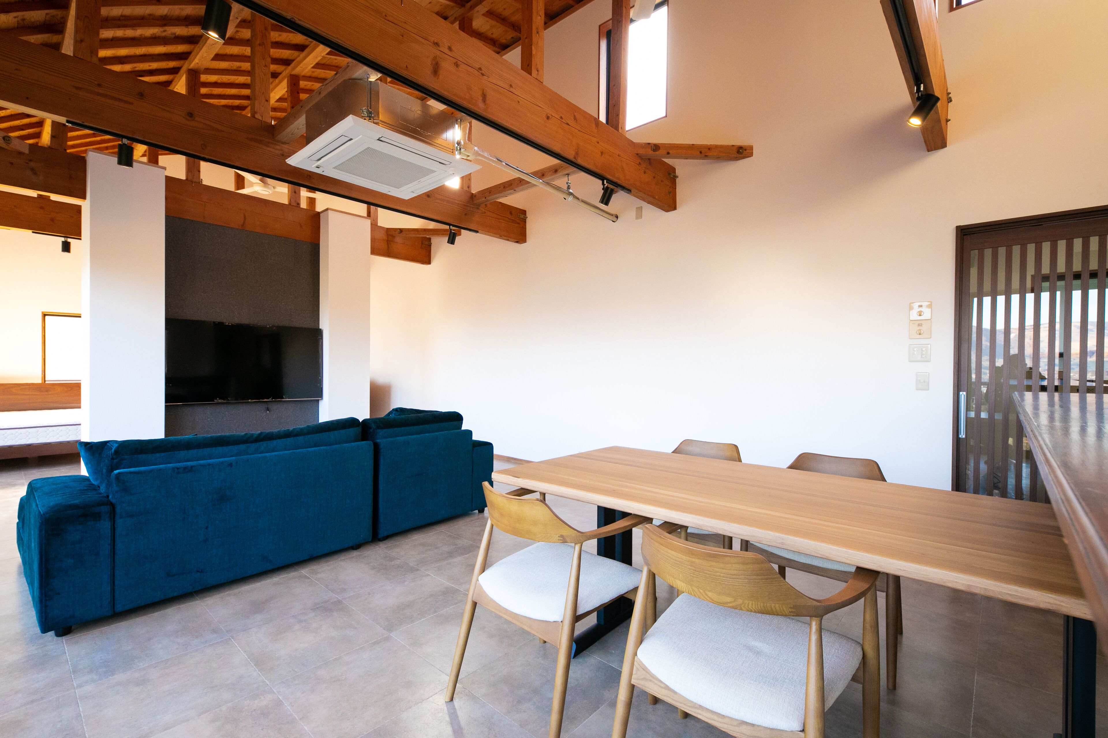 A modern living room with a wooden ceiling, a blue sofa, a wooden dining table with chairs, and a large flat-screen TV.