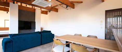 A modern living room with a wooden ceiling, a blue sofa, a wooden dining table with chairs, and a large flat-screen TV.